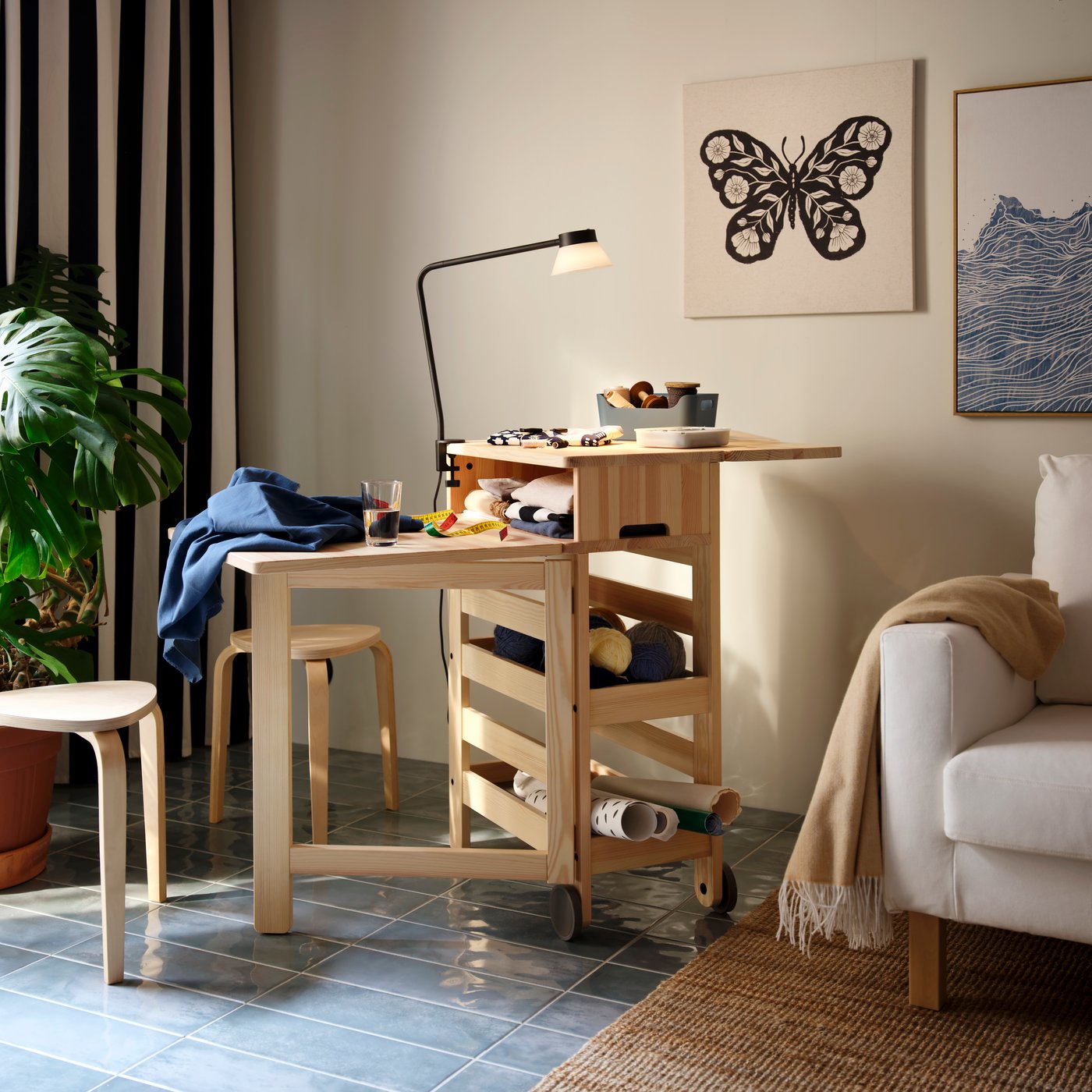 A sunlit living room with a pine RESARÖ mobile drop-leaf table, placed near an armchair and with textile accessories on top.