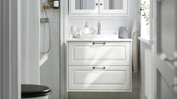 ホワイトのHEMNES/ヘムネス＆RÄTTVIKEN/レットヴィーケン 洗面台（引き出し×2）の上にLINDBYN/リンドビーン ミラーが掛かっている。隣にFRIHULT/フリフルト ウォールランプ。