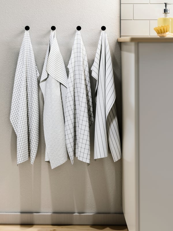 Four patterned, white-and-dark-grey RINNIG tea towels hang side-by-side on individual knobs by the end of a kitchen worktop.
