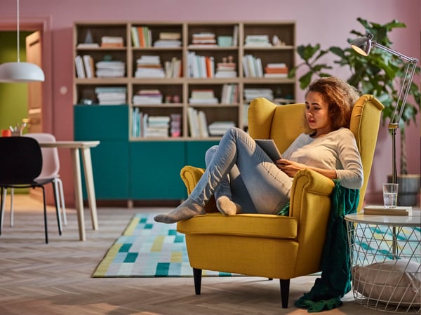 A young woman, curled up reading in a yellow STRANDMON armchair.