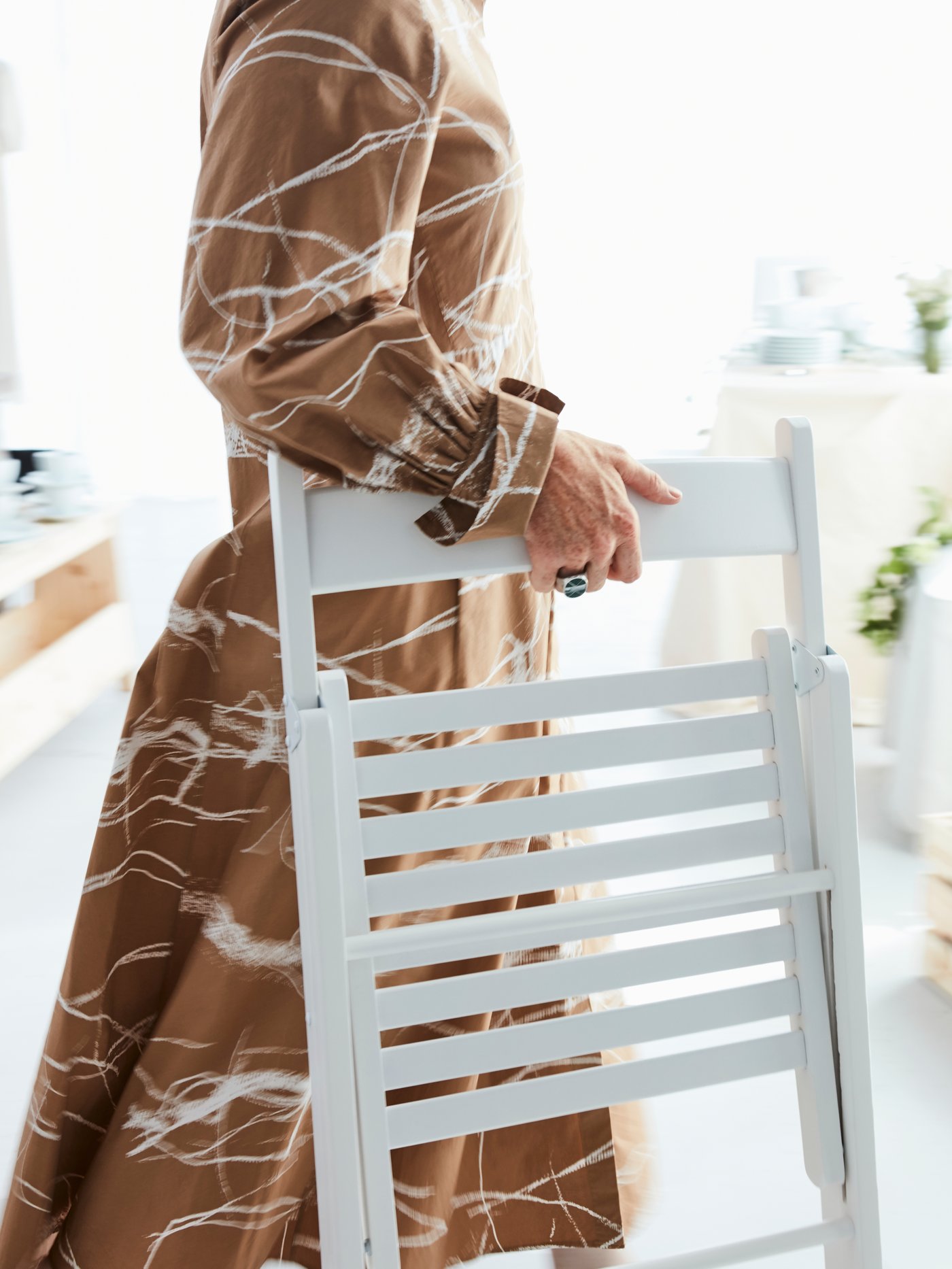 Dans un décor blanc, une femme vêtue d’une robe brune à motifs transporte une chaise pliante TERJE en bois blanc.