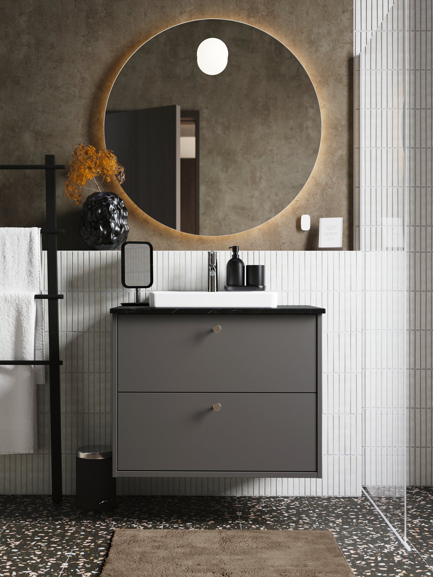 A matte, dark grey HÄVBACK wash-stand with two drawers and small knobs is in a bathroom below a round mirror lit from behind.