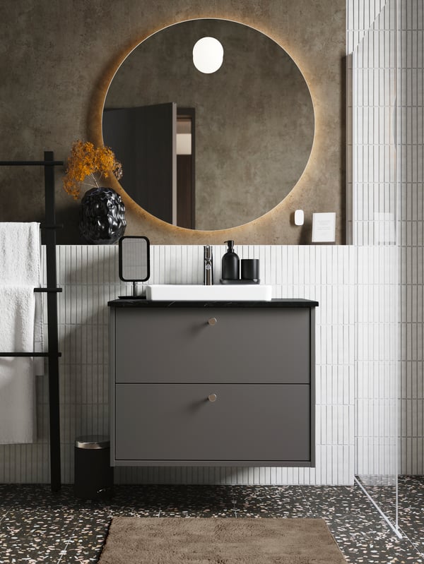 A matte, dark grey HAVBÄCK wash-stand with two drawers and small knobs is in a bathroom below a round mirror lit from behind.