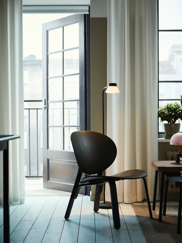 A SAMBORD floor/reading lamp stands beside a black stained oak veneer FRÖSET easy chair next to an open balcony door.