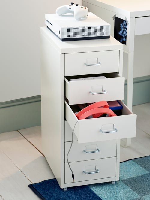 Workspace with white walls, white drawer unit on castors, white desk, rug, headphones, console with joystick.