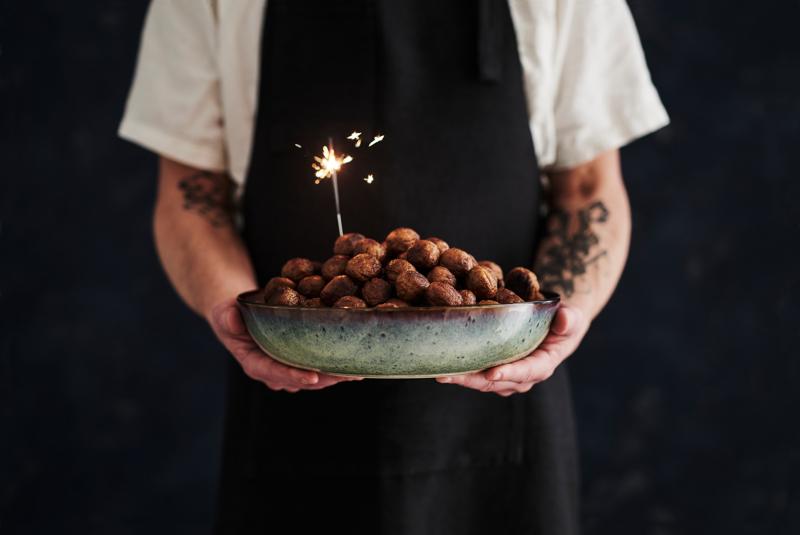 Personne avec un  tablier, un T-shirt blanc, des tatouages sur le bras droit, qui tient une assiette remplie de boulette et une bougie sur le dessus