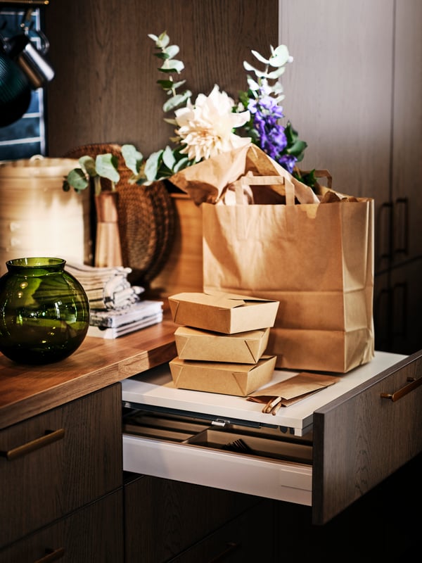 A green KONSTFULL vase on a worktop next to an open UTRUSTA pull-out work surface with a brown paper bag on top.