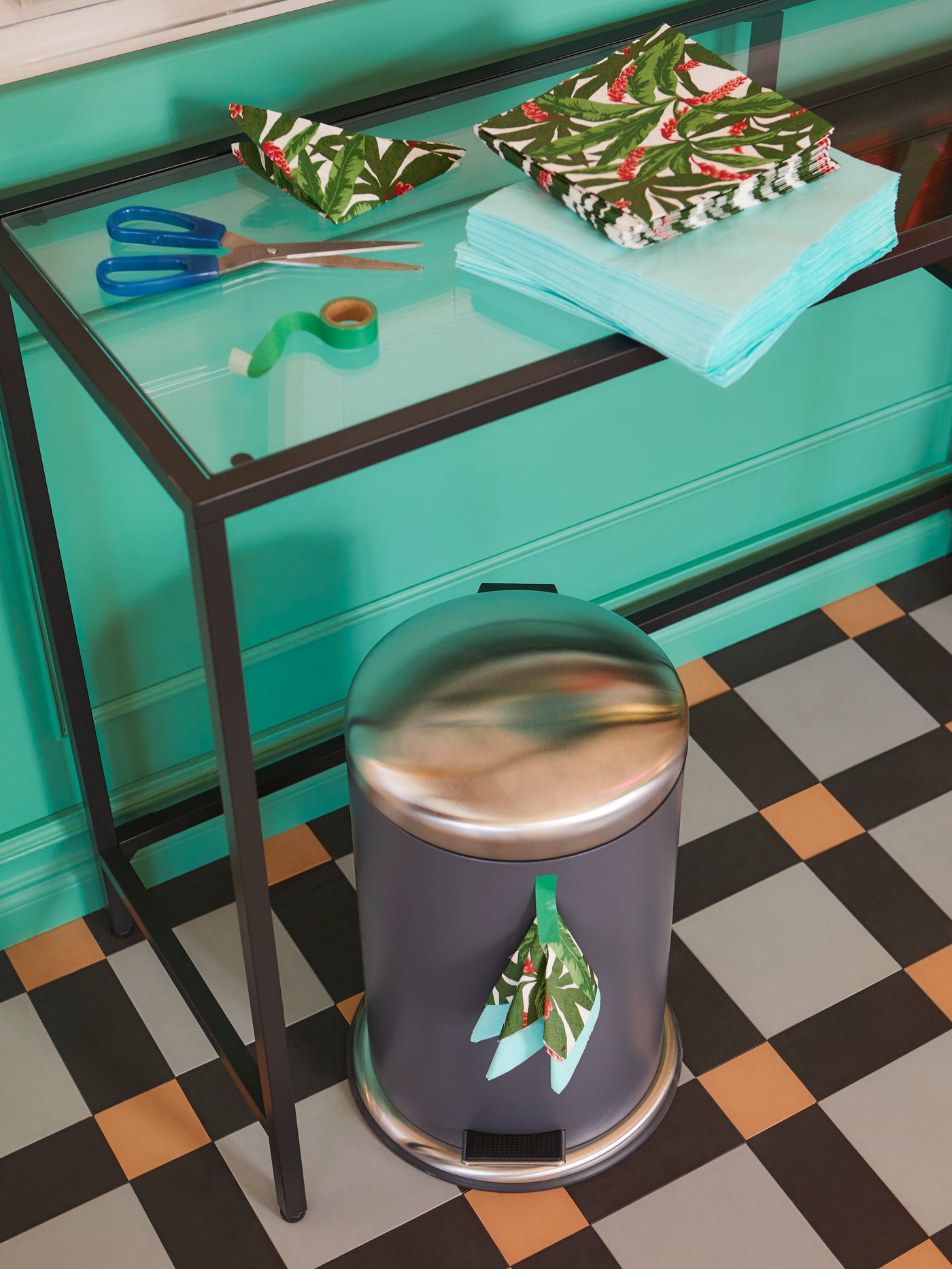 A VITTSJÖ laptop table placed along a wall, piles of FANTASTISK paper napkins on it and a MJÖSA pedal bin below.