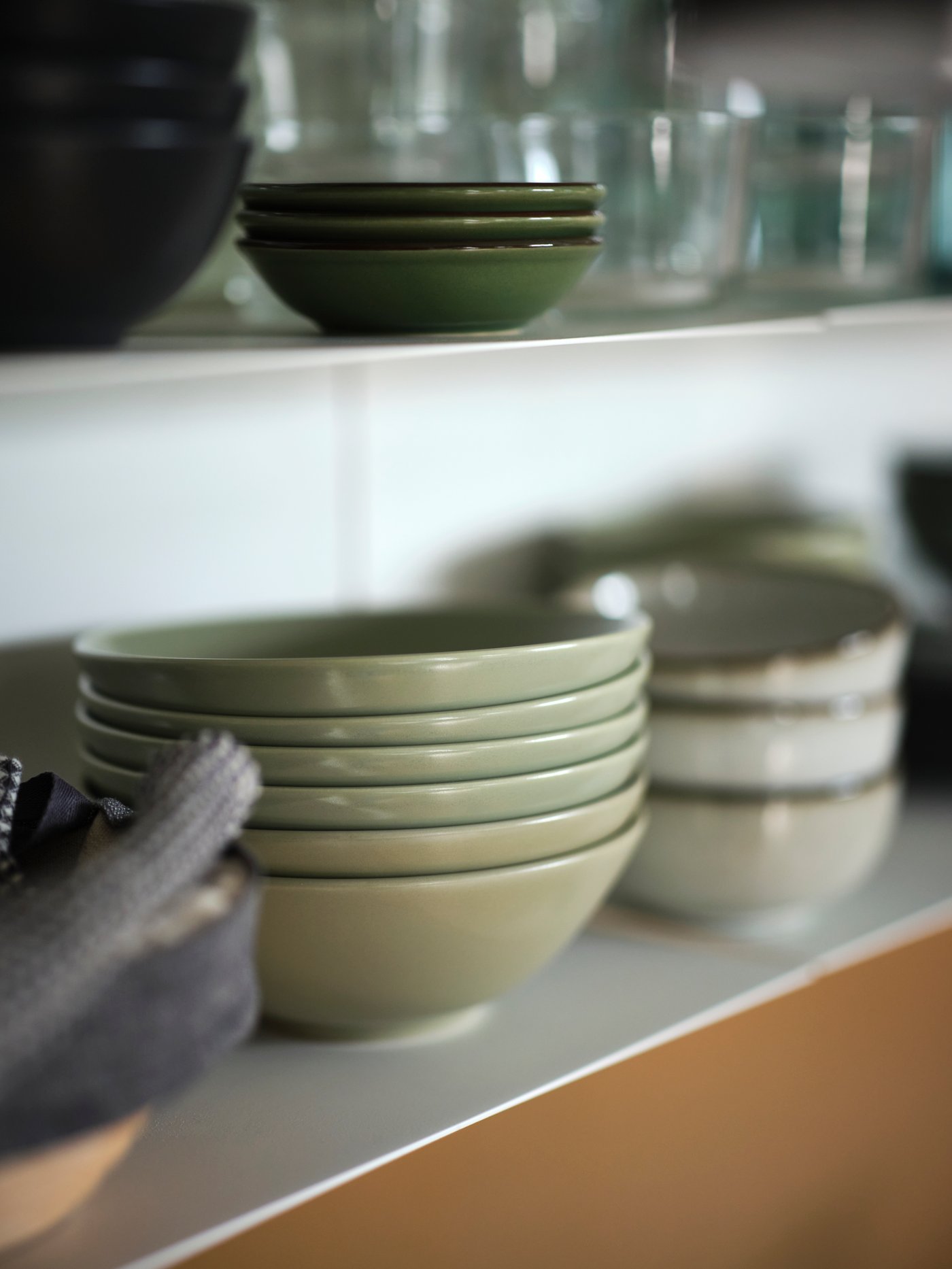 Shallow white shelves holding an assortment of crockery, including a stack of six matt green FÄRGKLAR bowls.