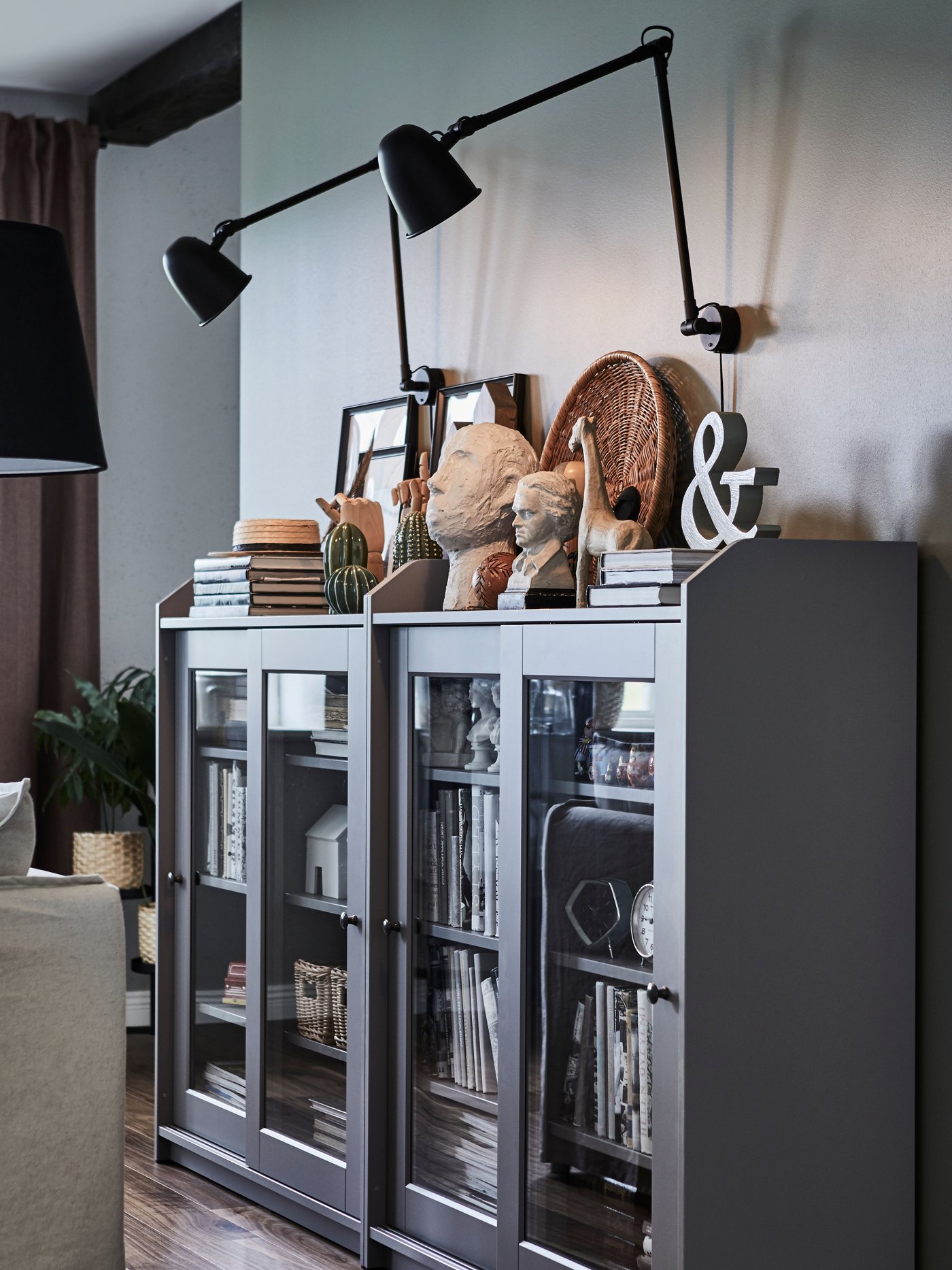 Two SKURUP work/wall lamps are attached to a wall above two grey HAUGA glass-door cabinets containing many items.