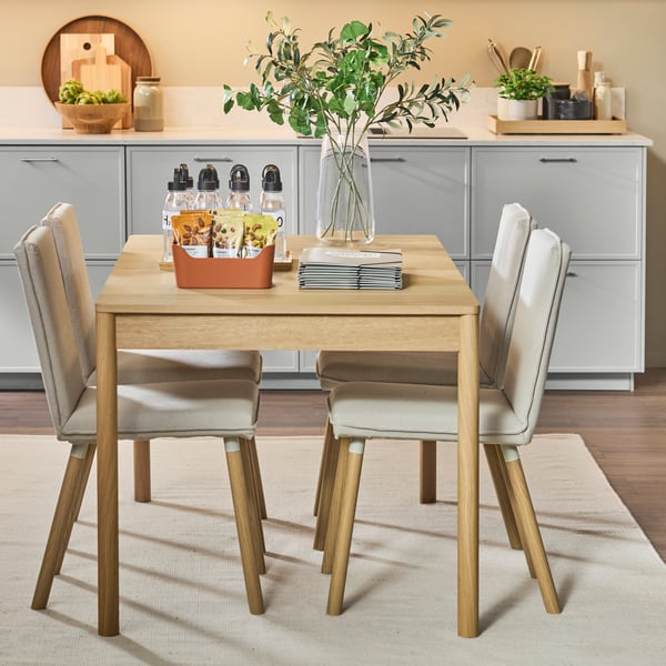 Dining table with chairs in a modern kitchen, vase of greenery and drinks on table, cabinets and decor arranged in background.