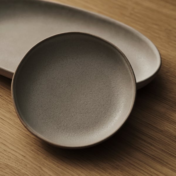A grey STOCKHOLM 2025 serving plate set of 2, with one round and one oval serving plate, on an oak veneer surface.