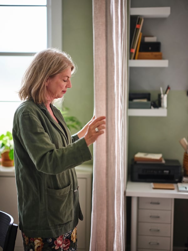 A woman closing a room-dividing fabric curtain, separating a home office with LACK wall shelves from another area.