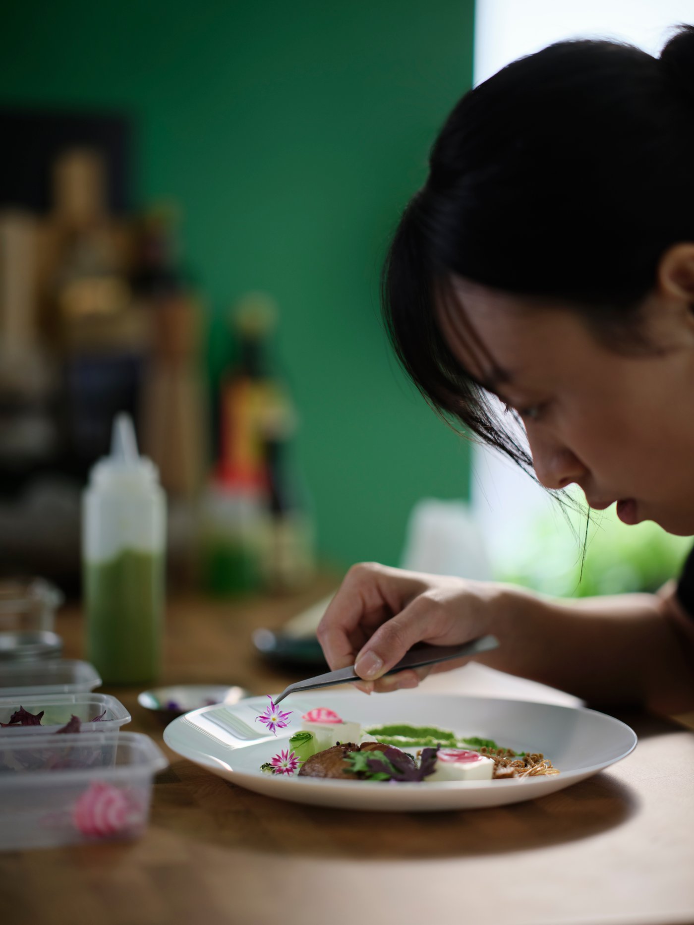 A person is carefully plating food on a white OFTAST deep plate with fresh ingredients and vibrant garnishes.