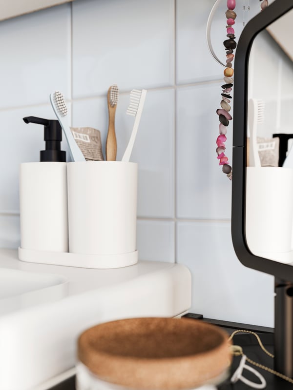 A white STORAVAN 3-piece bathroom set with toothbrushes, placed on a vanity and next to a black LINDBYN table mirror.