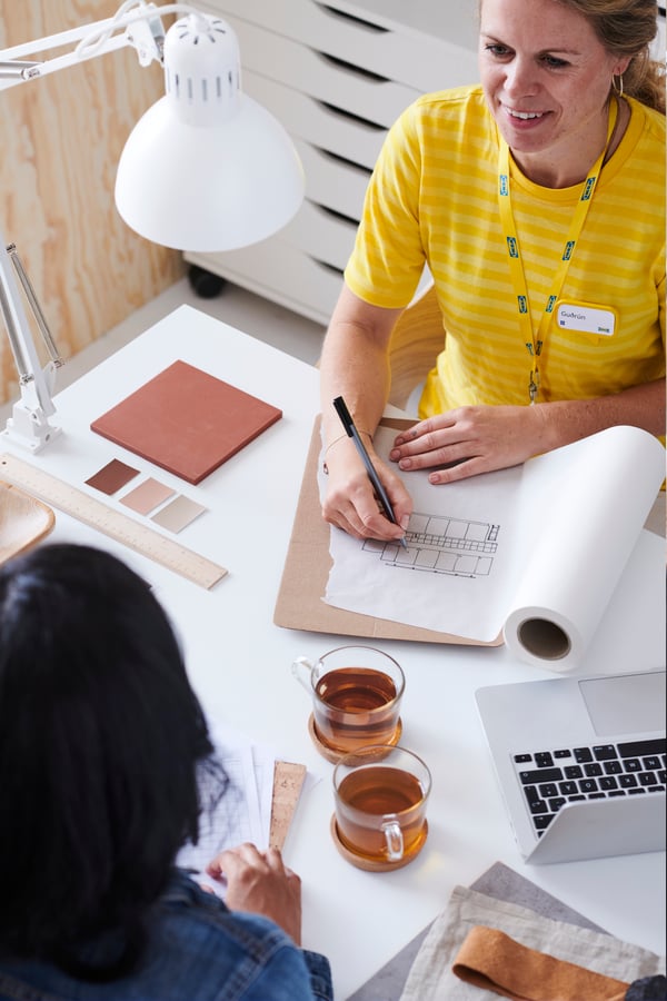 Deux personnes discutent d’un plan de construction à une table avec des échantillons de couleurs, un ordinateur portable, des tasses de thé et un dessin enroulé ; Une personne porte une chemise à rayures jaunes.