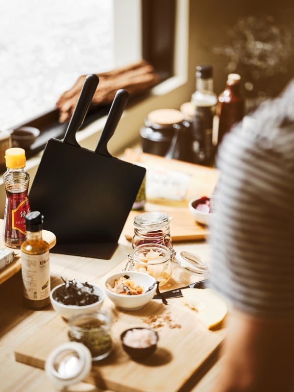 A worktop with a black JÄMFÖRA knife block with knives, an APTITLIG chopping board, KORKEN jars, and white bowls.
