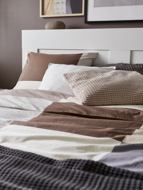 A white bed with headboard, brown patterned quilt cover and pillow cases, grey throw and cushions in white, grey and beige.