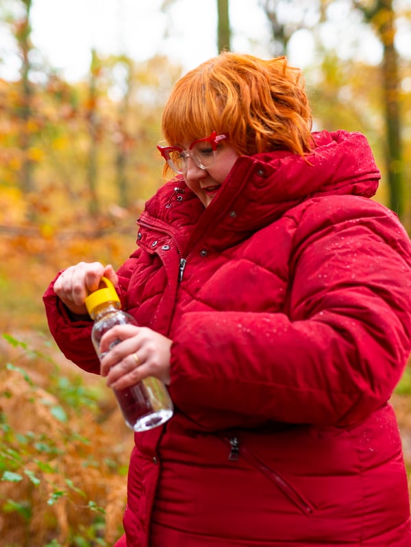 A woman in a forest is opening a FORMSKÖN water bottle. It's autumn and she is dressed in a warm coat.