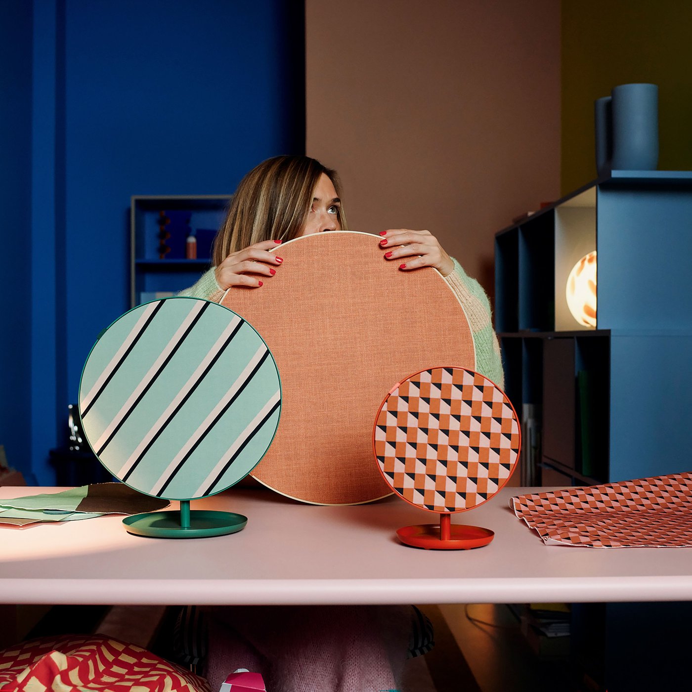 Tre runda högtalare i olika storlekar och mönster står på ett rosa bord. Den största panelen är enfärgad i orange, den mellanstora har diagonala svarta och vita ränder på turkos bakgrund, och den minsta har ett geometriskt mönster i orange och rosa. I bakgrunden syns hyllor och en blå vägg.