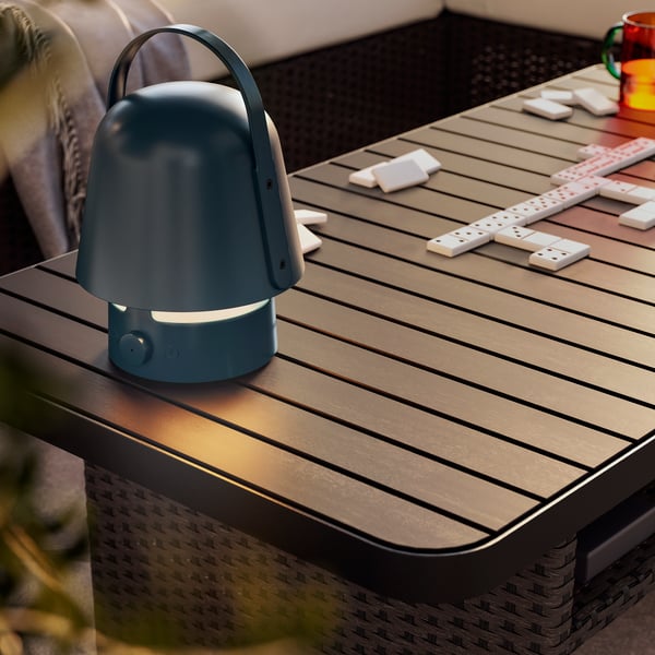A dark blue VAPPEBY bluetooth speaker lamp for outdoor use on a dark gray VITTSKÄR coffee table. Afternoon light outdoors.