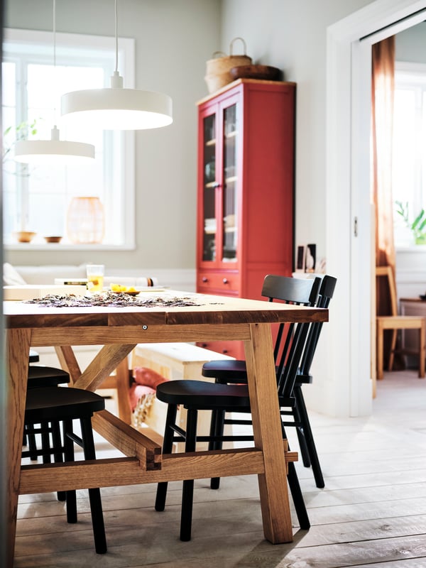 Two lit, white NYMÅNE pendant lamps above an oak MÖCKELBY table surrounded by black NORRARYD chairs.