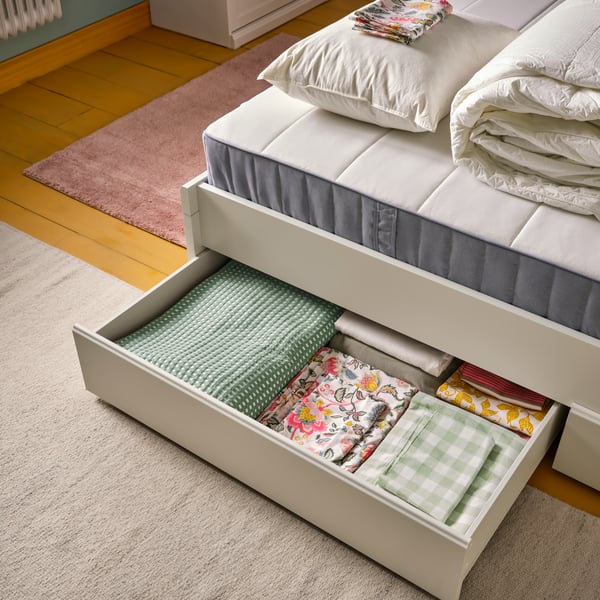 A bedroom with an off-white FÅGELFJÄLLET bed frame with four storage boxes. An open box is filled with neatly folded bedding.