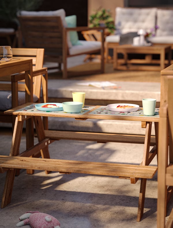 A sunny patio dining area with two tables and a RESÖ children’s picnic table with pastel coloured plates and mugs in between.