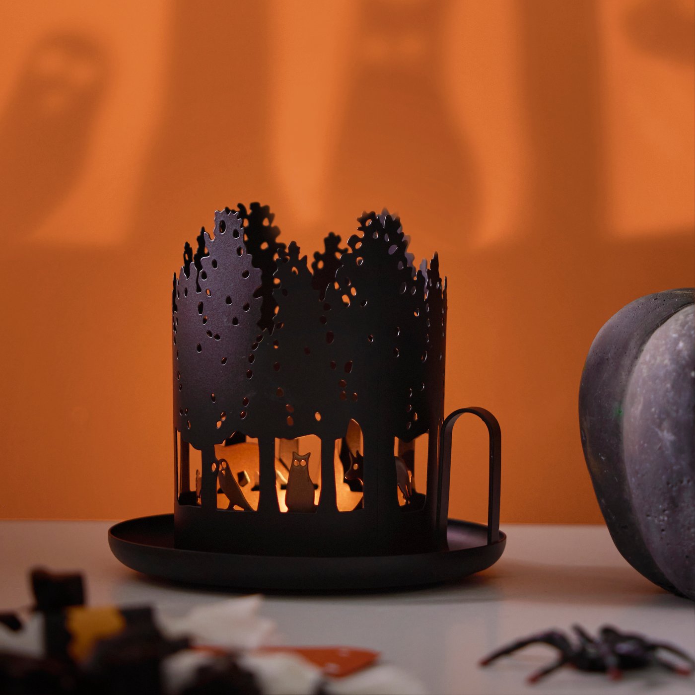 حامل شمعة صغيرة HÖSTAGILLE مزين بنقشات تجسد شكل حيوان وغابة، ينعكس ظلها على حائط بلون برتقالي.