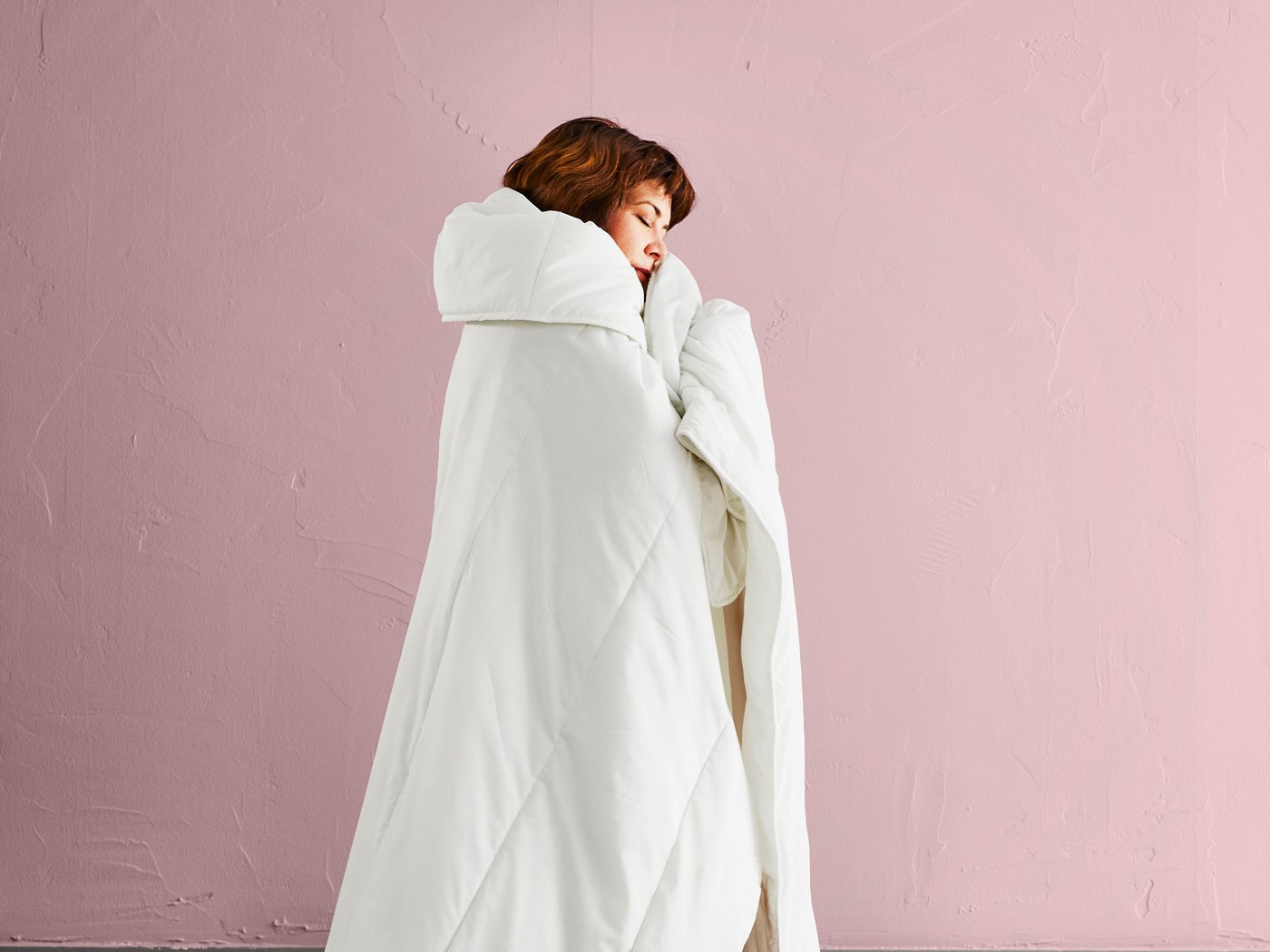 Woman wrapped up in a white SMÅSPORRE duvet against a pink wall.