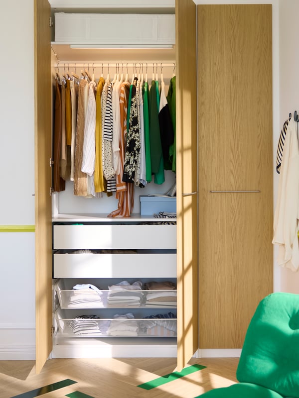PAX built-in wardrobe with STORKLINTA oak effect door, with one side of the wardrobe open to show integrated lighting, a clothes rail with BUMERANG hangers and SKUBB clothe storage boxes on the top shelf. KOMPLEMENT drawers and mesh baskets are also placed at the bottom half of the wardrobe. 