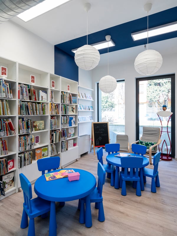 Area della Biblioteca comunale di Brescia Roncadelle allestita con tavolini e sedie MAMMUT di colore blu. La sala si affaccia su un’ampia vetrata da cui si vedono le librerie BILLY bianche disposte lungo le pareti e le lampade a sospensioni REGOLIT.