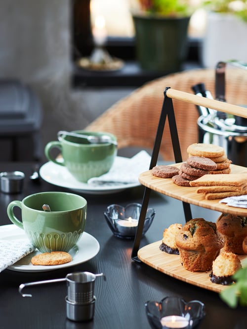 Gros plan sur une table sur laquelle reposent des tasses, des bougies chauffe-plat ainsi qu'un plateau étagé FULLSPÄCKAD en bambou et acier peint noir sur lequel se trouvent un assortiment de pâtisseries.