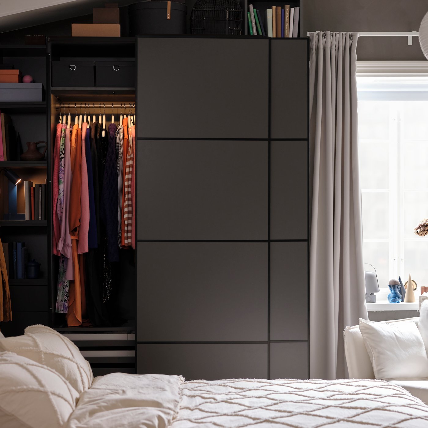 Achter een bed staat een donkergrijze PAX kleerkast met een schuifdeur die op een kier staat, zodat kleding zichtbaar is die in de kast aan een kledingroede hangt.