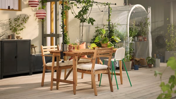 Terrasse lumineuse avec table d’extérieur ASKHOLMEN en acacia, chaises d’extérieur avec accoudoirs et de nombreuses plantes.