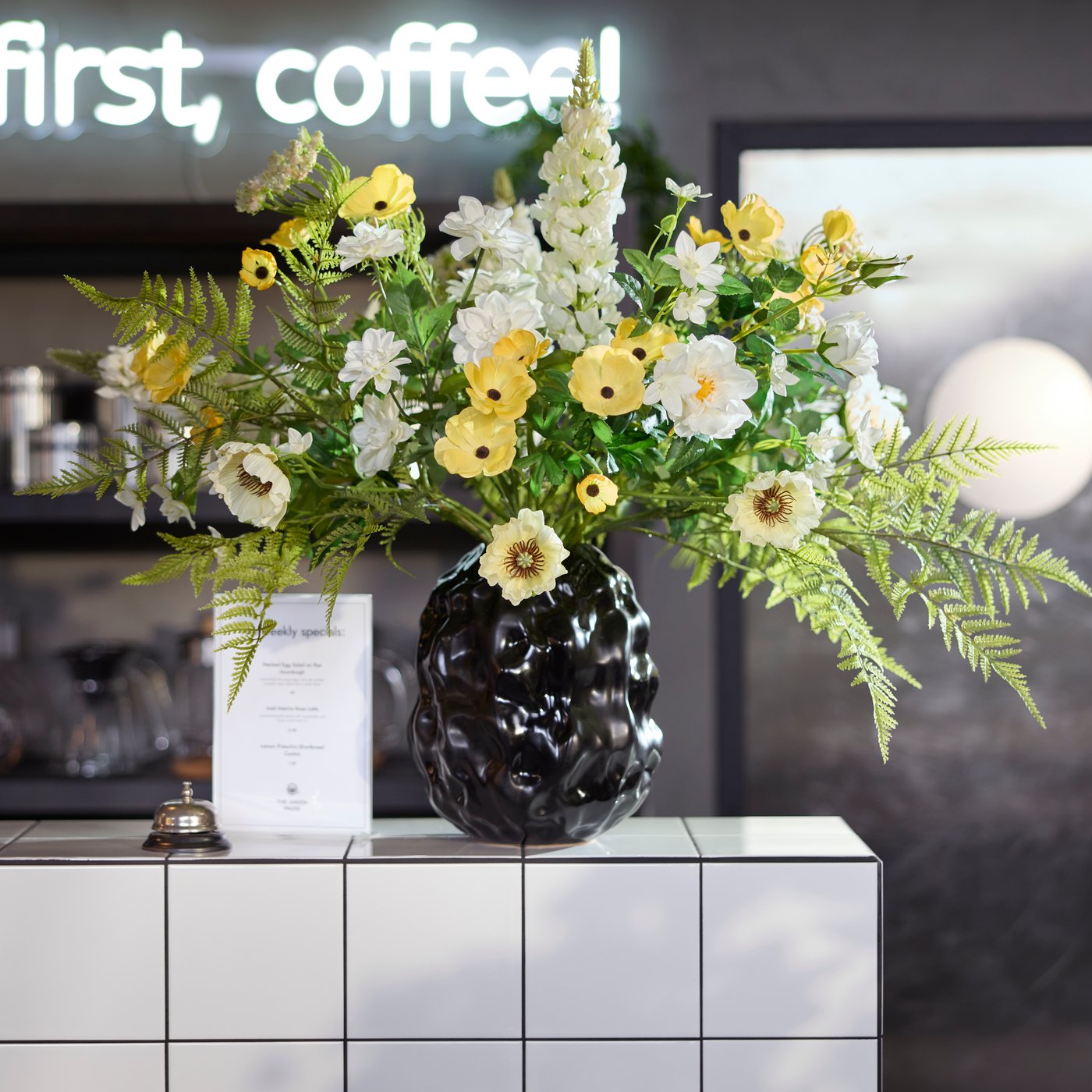 A café counter by a lighting sign shows a black STOCKHOLM 2025 vase filled with colourful flowers alongside a menu and bell.