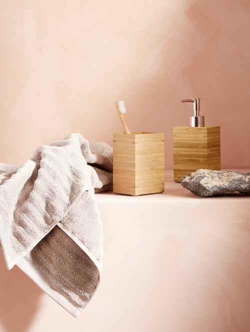 Salle de bains avec mur rose, porte-brosses à dents en bambou carré et distributeur de savon en bambou, avec une serviette éponge beige.