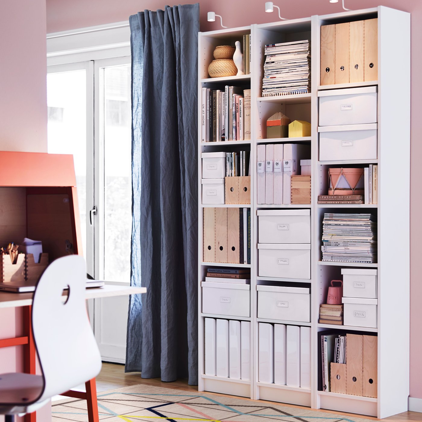 Three white BILLY bookcases with open shelves filled with boxes, organizers, and books against a pink wall in a bright room.