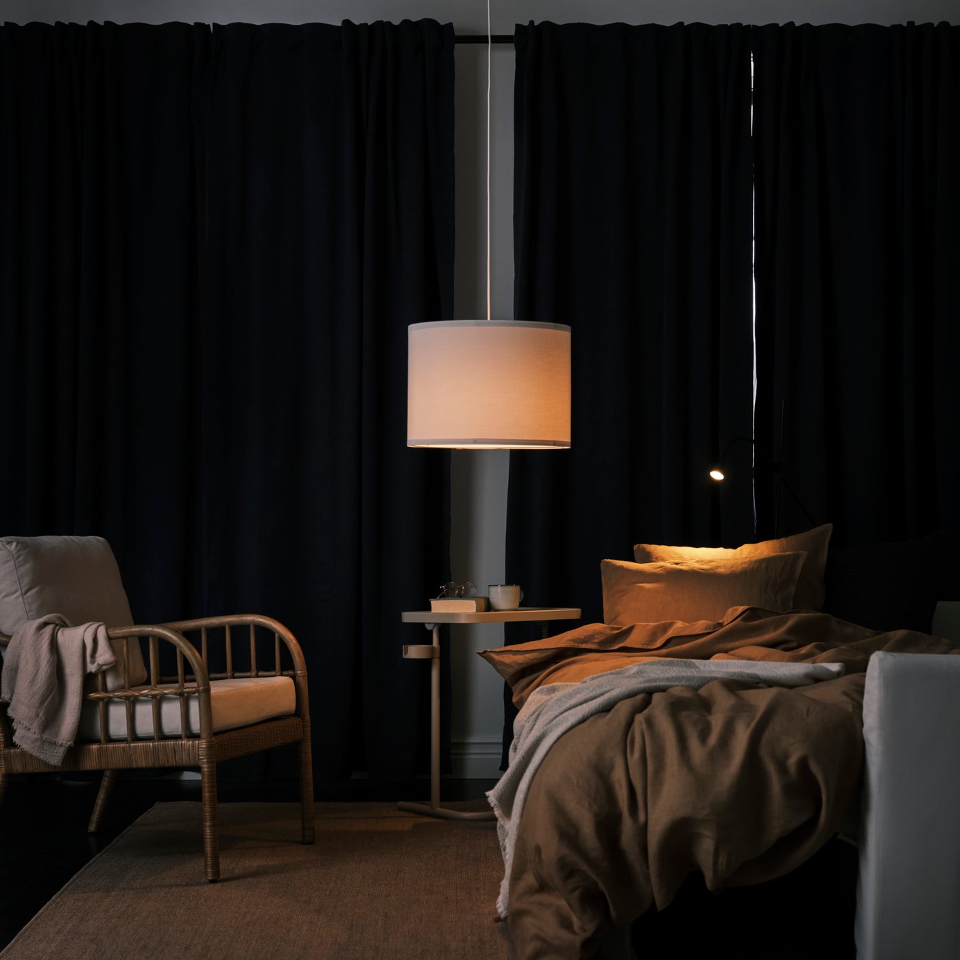 Ett mörkt, stämningsfullt sovrum med bäddad säng, fåtölj och två lampor som lyser varmt mot mörka gardiner. Ett sidobord står mellan fåtöljen och sängen.
