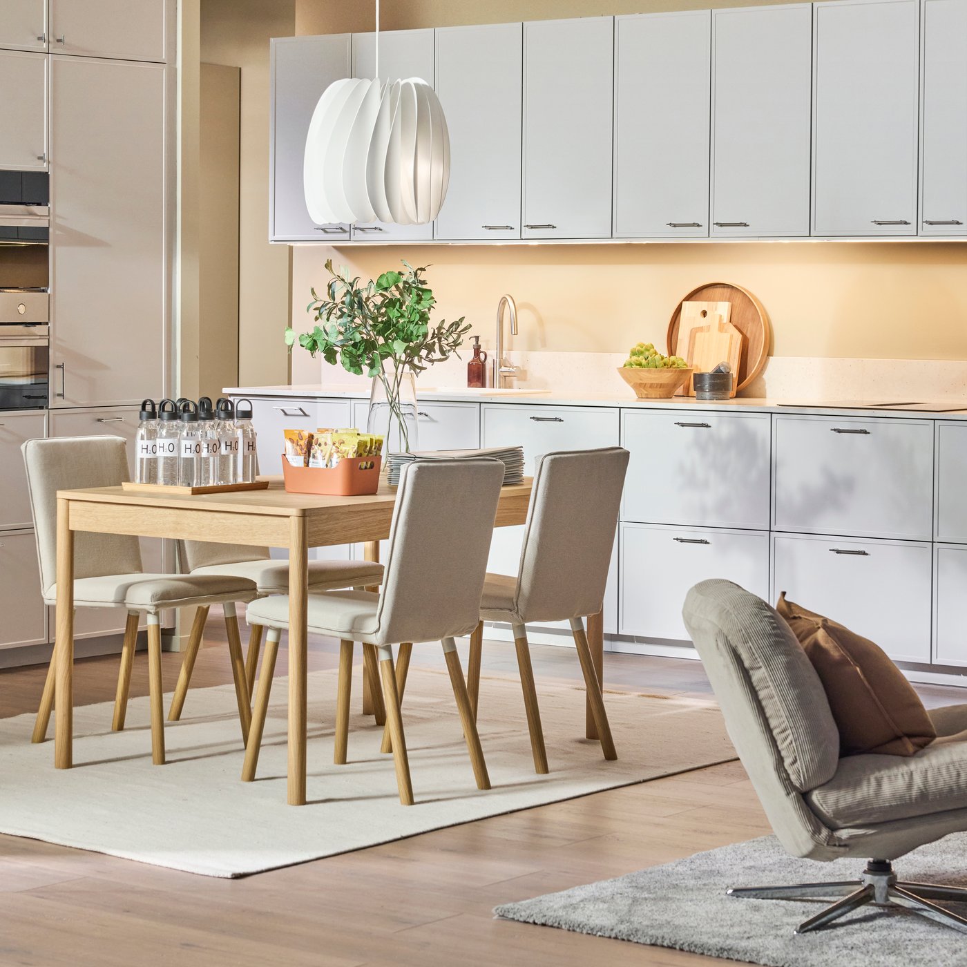 Ljus, modern köksinredning med vita skåp, matbord och stolar, pendellampa, växter och prydligt arrangerade föremål.