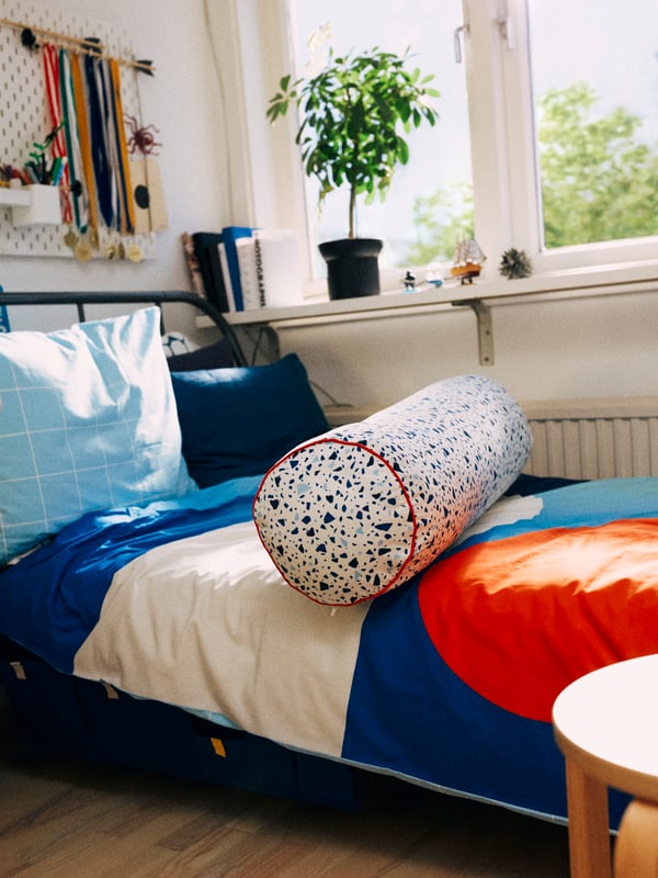 A bedroom with a KOPARDAL bed by a window with a multicolored BRUNKRISSLA quilt and pillowcase and a MÖJLIGHET cushion.