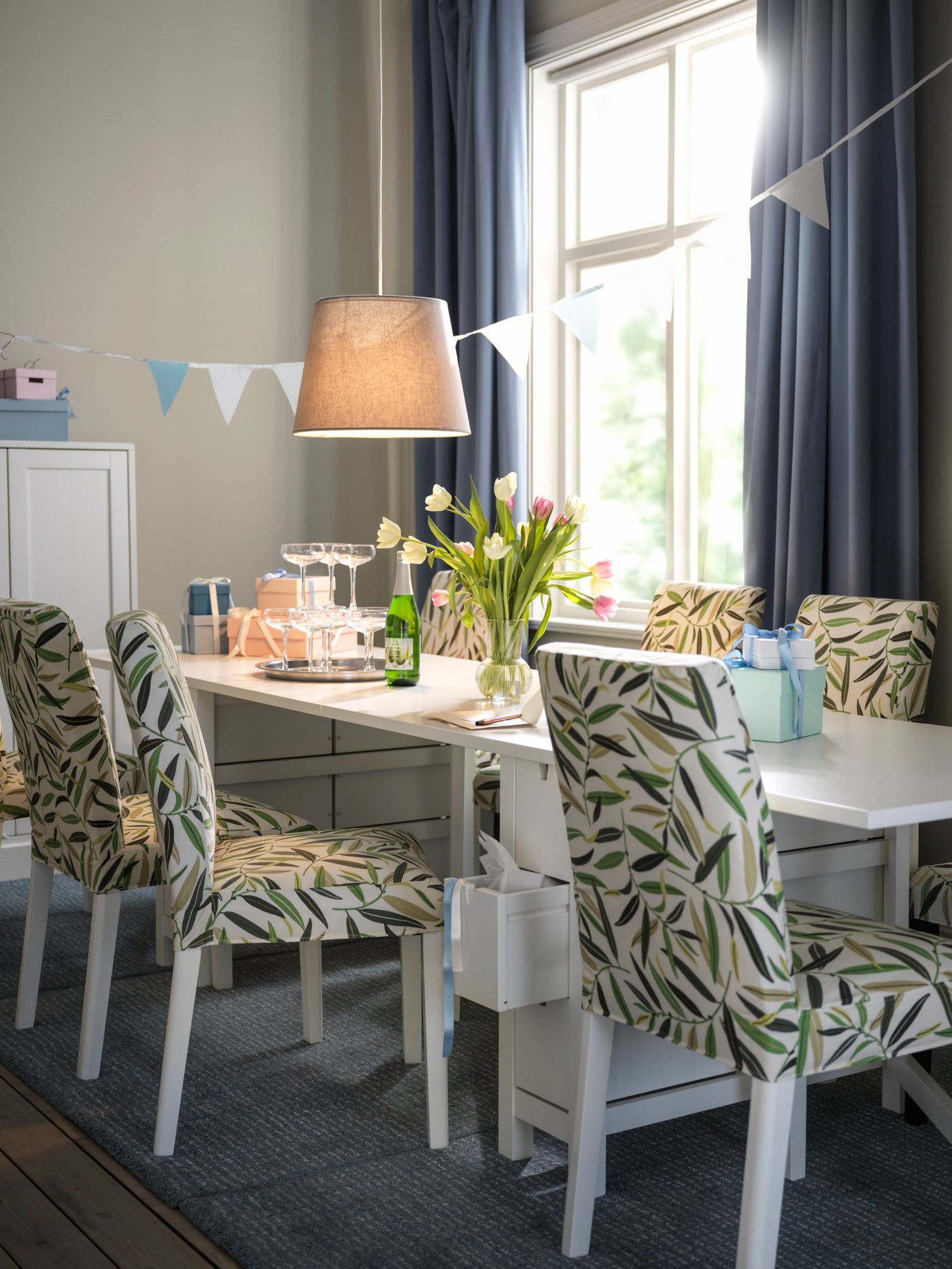 Comedor con una mesa plegable abierta, varias sillas blancas, alfombra de pelo corto, pantalla de lámpara colgante y cortinas semiopacas.