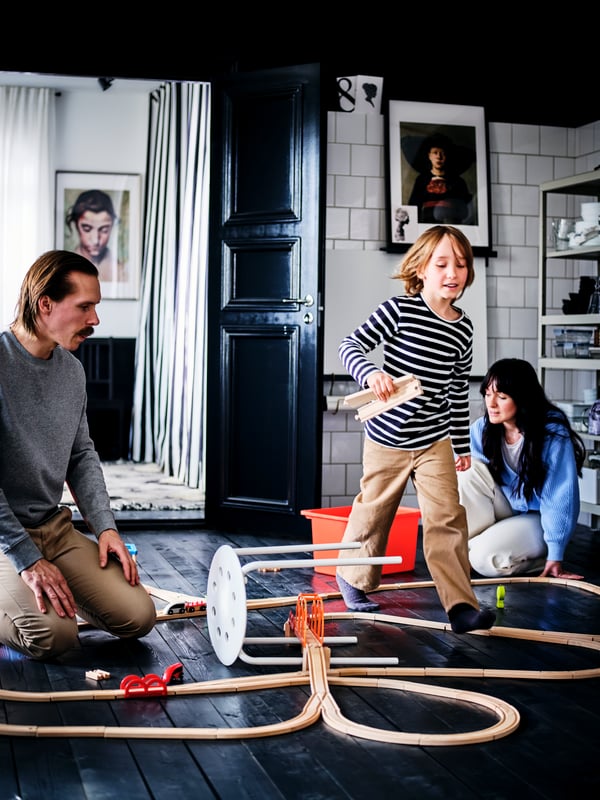 Parents and a child share a play moment over a LILLABO rail system, including a white MARIUS stool, on a dark wood floor.
