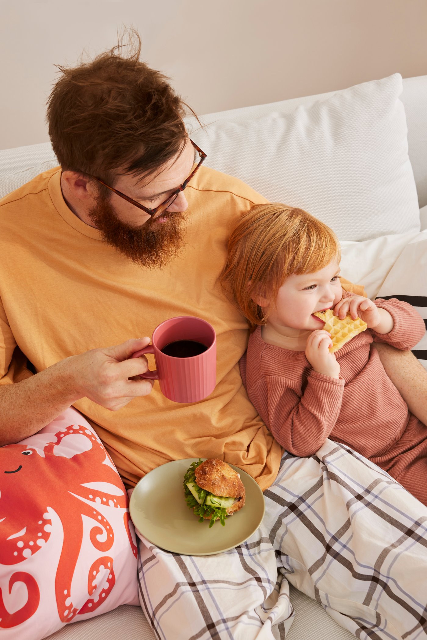 Ein Vater mit einem Kaffee und seinem Kind in seinem Arm, das gerade eine Waffel isst.