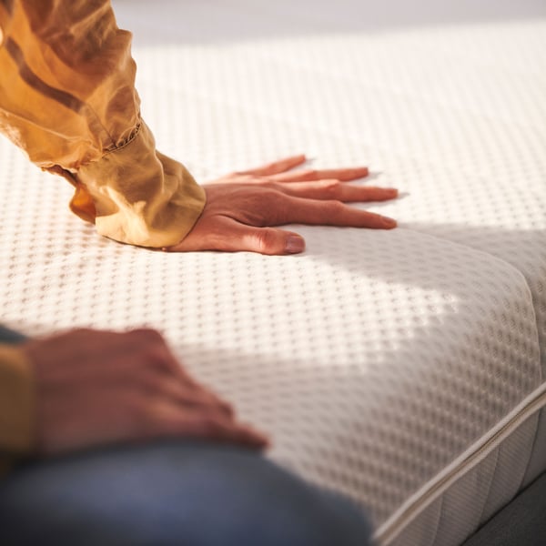 Une personne portant un chemisier jaune est assise sur le coin de deux matelas en mousse ÅKREHAMN blancs. Une main touche la surface.