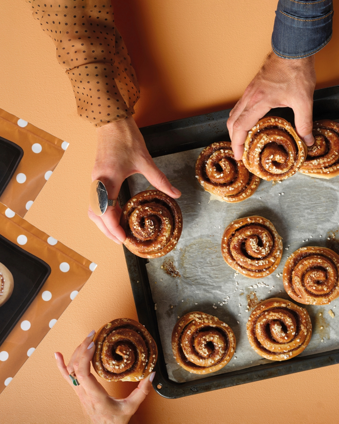 Cinnamon buns on a baking tray, held by three people, orange table in the background.
