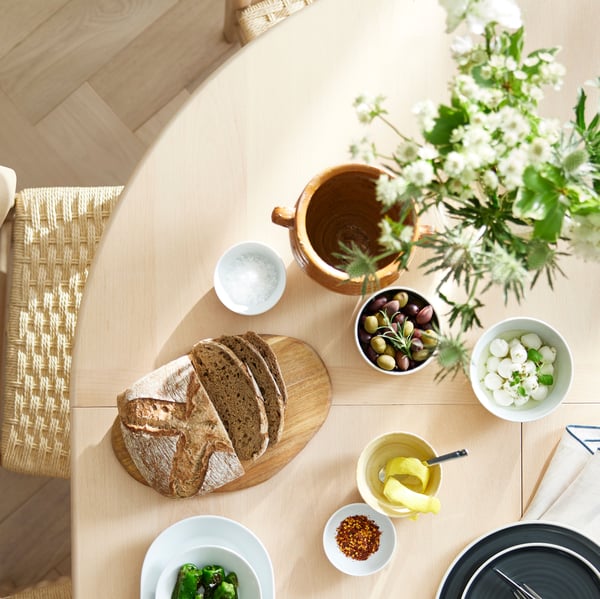 Part of the top of a light beech/veneer SKANSNÄS round extendable table with various types of food and some flowers on it.