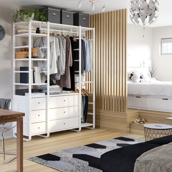 Ein weißer, offener Kleiderschrank der ELVARLI Serie neben einem Bett in einem hellen Raum.
