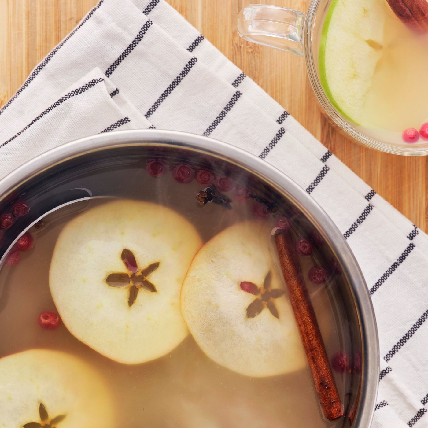 À côté d’un mug, faitout contenant du thé vert FLÖJT chaud, des tranches de pomme et des épices, sur un tissu rayé.