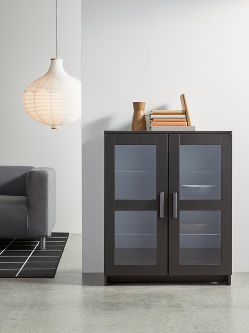 A glass/black BRIMNES cabinet with doors has decorative bowls inside and books on top. It is in a display-style living room.